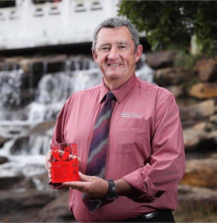 2022 Australia Day Ambassador for Inverell Shire, Chief Inspector Bob Fitzgerald pictured in a red button up shirt and patterned tie, holding the award for 2019 Volunteer of the Year. 
