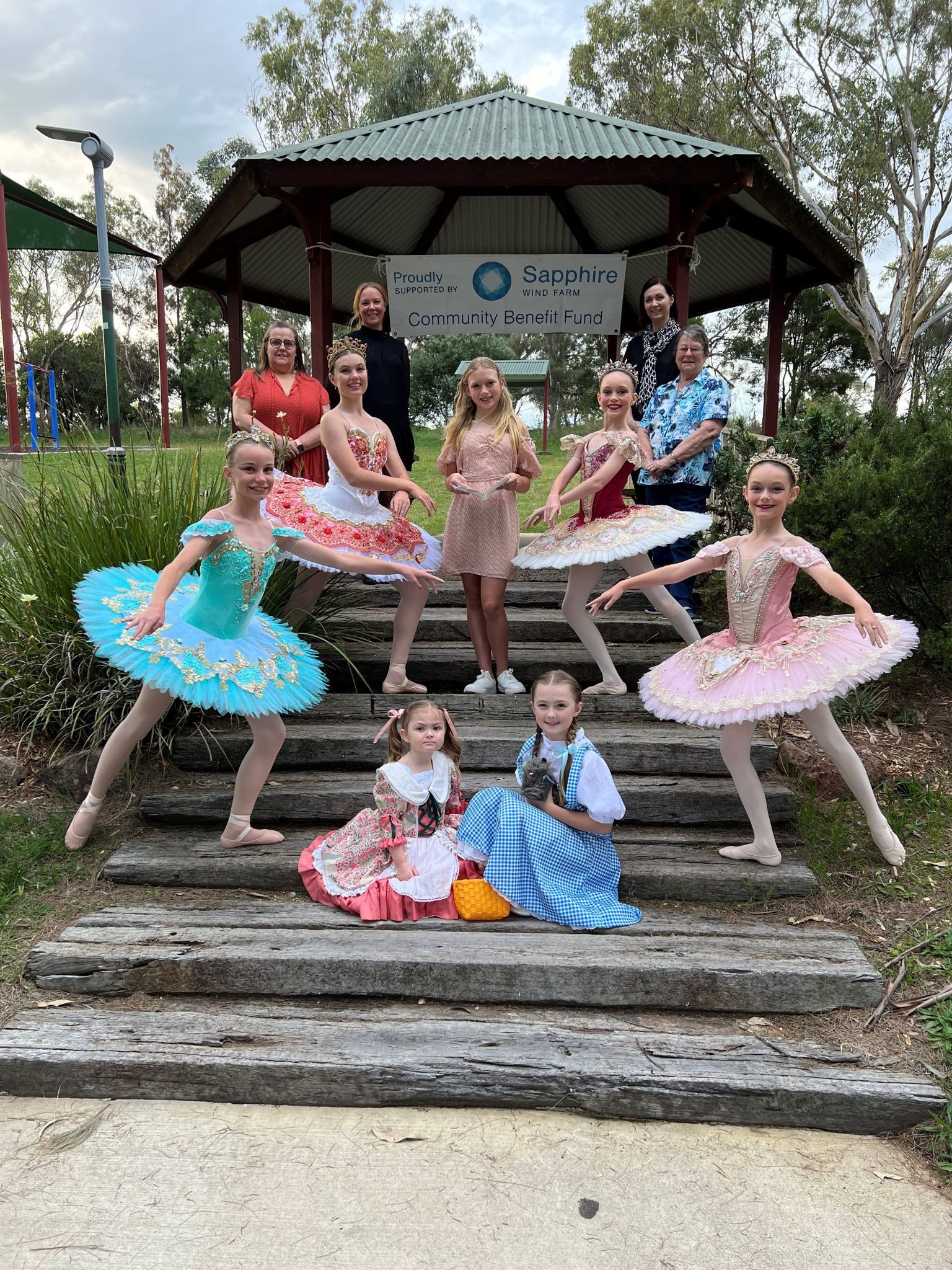 Eisteddfod performers standing in full costume on tiered staircase at a Lake Inverell pergola, with Sapphire Wind Farm Banner stretched across the back. 